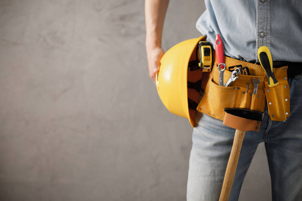 Worker man and tool belt holding construction helmet near wall. Male hand and tools for house room renovation. Home renovation concept - Fotoğraf, Görsel