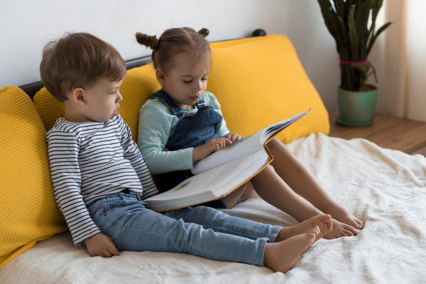 Two preshool, toddler children read large interesting book of fairy tales on yellow bed. Siblings little twins reader have fun, happy kid on quarantine at home. Friendship, family, education concept - Foto, Imagen