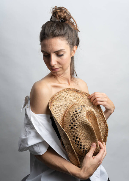 Model, brünett mit Strohhut posiert auf weißem Hintergrund im Studio. Sie trägt ein weißes Hemd, eine schwarze Frisur. - Foto, Bild