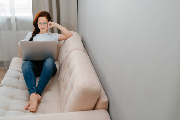 Young woman with glasses is sitting on the couch watching a movie on a laptop. - Фото, изображение