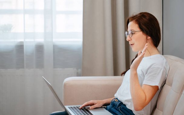 Woman in a white T-shirt at home on the couch communicates online via video help on a laptop.Concept of a freelancer. - Foto, Imagem