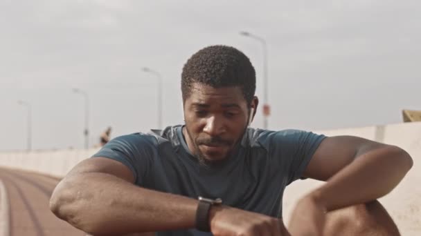 Slowmo médio tiro de jovem atlético afro-americano limpando suor da testa sentado na pista de corrida ao ar livre na manhã ensolarada, descansando depois de correr - Filmagem, Vídeo
