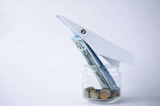 Paper plane with image of dogecoin aims upward, leaning on glass jar with coins and dollars. Cryptocurrency. - Foto, afbeelding