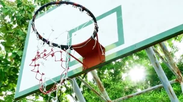 Μπάσκετ στεφάνι σε υπαίθριο δημόσιο γήπεδο στο φως του ήλιου λάμπει μέσα από πράσινο φόντο δέντρα. - Πλάνα, βίντεο