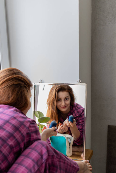 Young red-haired girl sits the windowsill in front of mirror and puts on makeup. Concept of caring for your appearance. Home interior - Foto, imagen