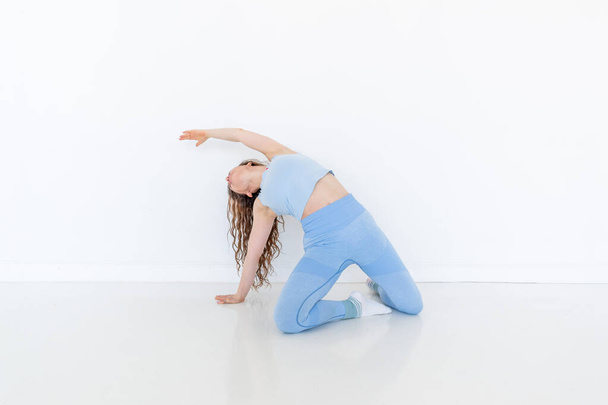 Young diverse woman practices yoga, doing body stretching exercise. Sitting on the floor. White studio loft. Sports and wellness concept. Copy space - Foto, afbeelding