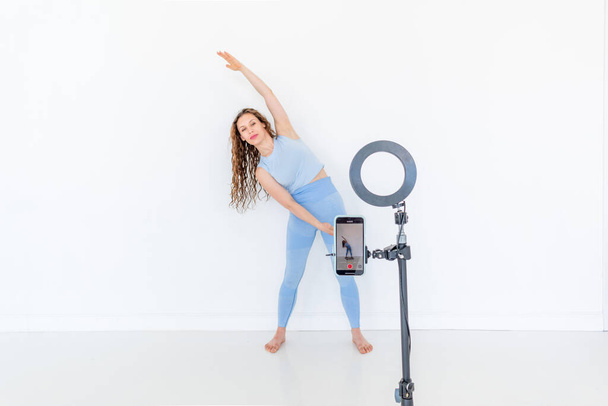 Young diverse woman is recording a dance instructional video on her phone. White studio loft. Online training concept. Copy space - Фото, изображение