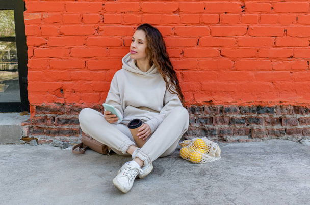Adult woman sits on the floor with a phone in her hands and a thermo mug with a drink. Wellness concept. - Foto, Imagen