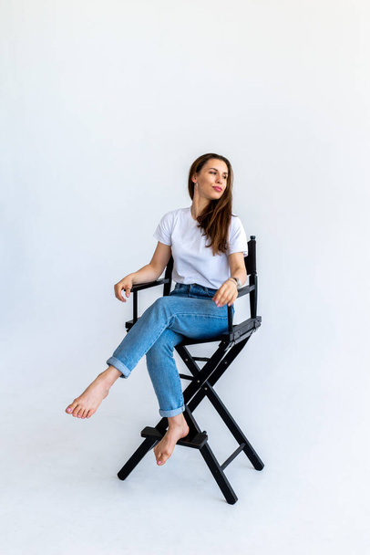 Adult woman sits on a filmmaker's chair in blue jeans and a white T-shirt. Red hair in humans. White background - Foto, afbeelding