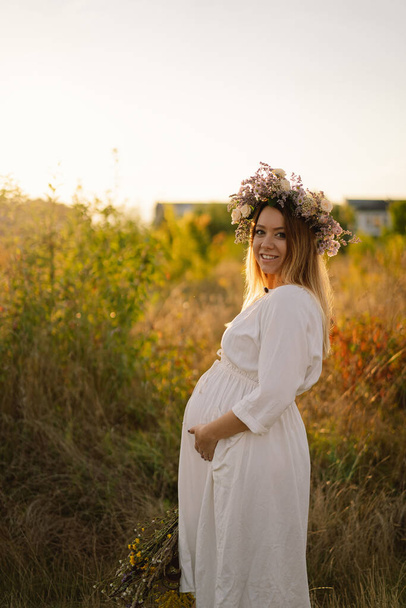 Portrait of a pregnant woman. A beautiful young pregnant woman in a white dress walks in the field. - Фото, зображення