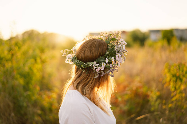 Όμορφη γυναίκα με στεφάνι από αγριολούλουδα. Στέκεται πίσω στον τομέα των λουλουδιών, τα χέρια στο πλάι - Φωτογραφία, εικόνα