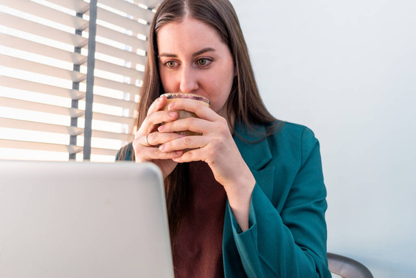 Young woman in suite is working on computer and while drinking coffee. Businesswoman is keeping social distancing and working remotely. New normal concept - Foto, Imagen