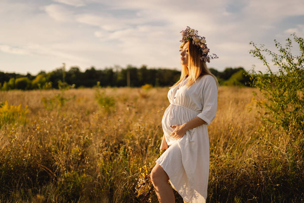 Portrait of a pregnant woman. A beautiful young pregnant woman in a white dress walks in the field. - Фото, изображение