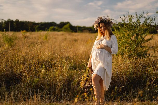 Портрет беременной женщины. Красивая молодая беременная женщина в белом платье гуляет по полю. - Фото, изображение