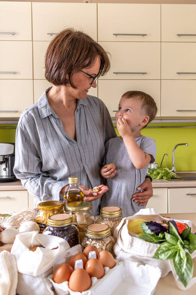 Adult mother feeds her son with nuts in the kitchen. Eco-friendly shopping in fabric bags and glass jars. Zero waste concept. - 写真・画像
