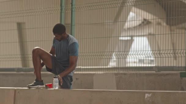 Genç Afro-Amerikan atletizm sporcusunun şehir köprüsü altında tek başına koşarken ayakkabılarının bağcıklarını bağladığı yavaş çekim. - Video, Çekim