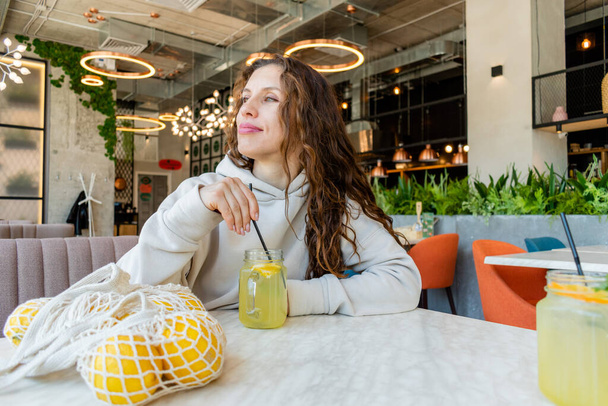 Diversity woman with red hair sits alone in cafe drinking lemonade from a glass jar. Refreshing drink in summer heat. Concept of wellness and proper nutrition. - Photo, image