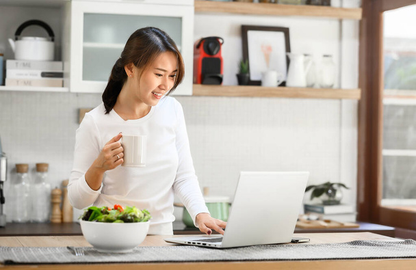 gelukkig aziatische vrouw werken thuis met behulp van laptop in keuken in tne ochtend - Foto, afbeelding