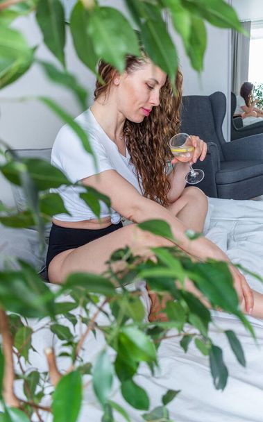 Young woman ginger hair sitting the bed in bedroom and drinks water with lemon. Fresh morning. Wellness and detox concept. Copy space - Photo, Image