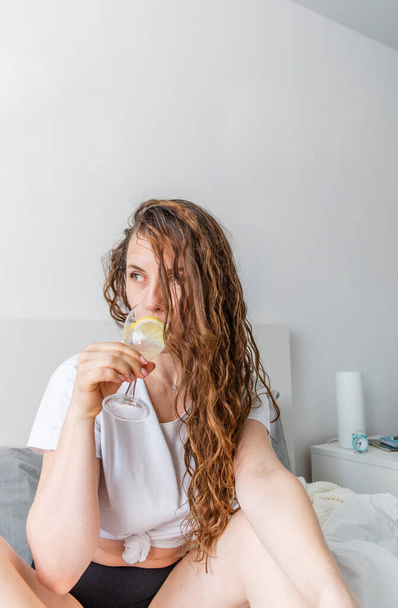Young woman ginger hair sitting the bed in bedroom and drinks water with lemon. Fresh morning. Wellness and detox concept. Copy space - Fotó, kép