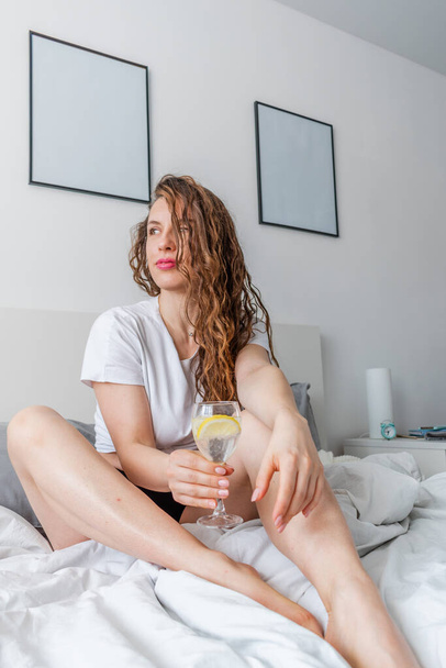 Young woman ginger hair sitting the bed in bedroom and drinks water with lemon. Fresh morning. Wellness and detox concept. Copy space - Φωτογραφία, εικόνα