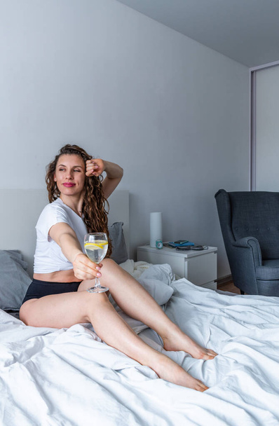 Young woman ginger hair sitting the bed in bedroom and drinks water with lemon. Fresh morning. Wellness and detox concept. Copy space - Фото, зображення