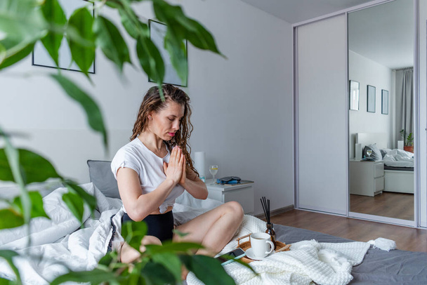 Young business woman meditates while sitting on the bed, after work. Girl listens calm music near telephone. Comfort zone and wellness concept. Yoga at home - Foto, Bild