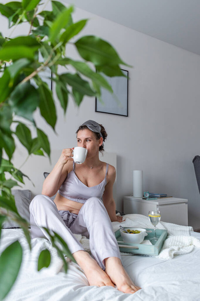 Young woman in silk pajamas has healthy breakfast while lying on bed in bedroom. Home wellbeing concept. Emotional health of a young woman - Zdjęcie, obraz