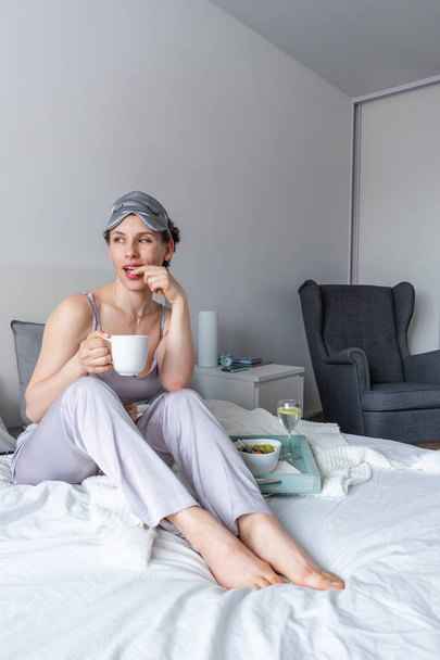 Young woman in silk pajamas has healthy breakfast while lying on bed in bedroom. Home wellbeing concept. Emotional health of a young woman - Photo, image