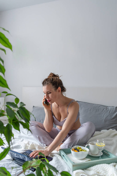 Young woman in comfortable lilac pajamas works on a computer and talks a cell phone while lying the bed in the bedroom. Business woman maintains a social distance works remotely. New normal concept - Foto, Bild
