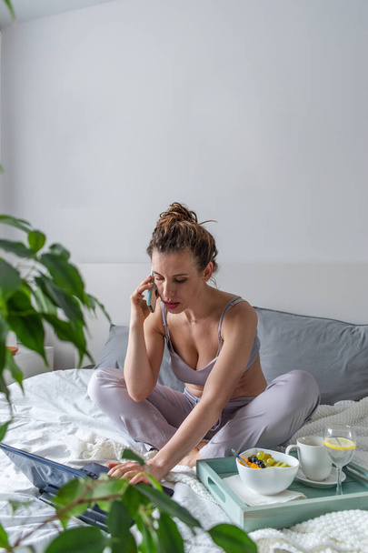 Young woman in comfortable lilac pajamas works on a computer and talks a cell phone while lying the bed in the bedroom. Business woman maintains a social distance works remotely. New normal concept - Фото, зображення