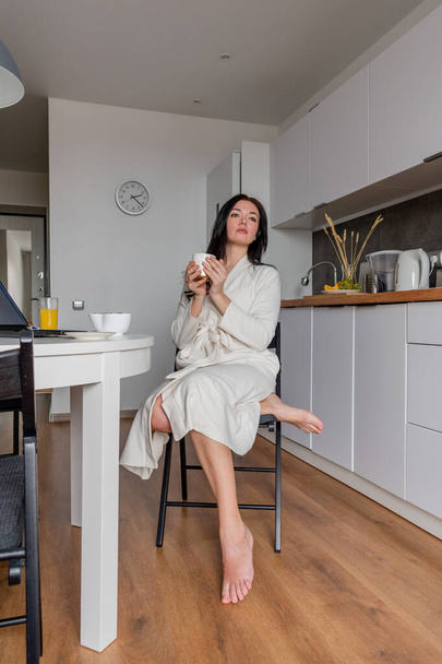 Young woman with black hair in white robe sits on chair the kitchen. Hands hold white mug with aromatic coffee. Wellness and healthy food concept - Foto, afbeelding