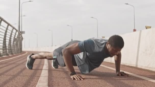 Langzame opname van atletische Afro-Amerikaanse man in sportkleding en draadloze hoofdtelefoon doen klappen push-ups training buiten op de racebaan op de zomerochtend - Video