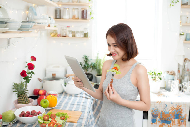 Szczęśliwy piękny asian kobieta przygotowuje zdrowe jedzenie w kuchni i czytaniu przepis za pomocą cyfrowego tabletu. - Zdjęcie, obraz