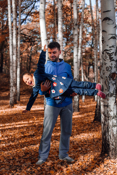 full length happy family concept. man with dark hair play and hold his little daughter in blue jeans coat at autumn forest background - Photo, Image