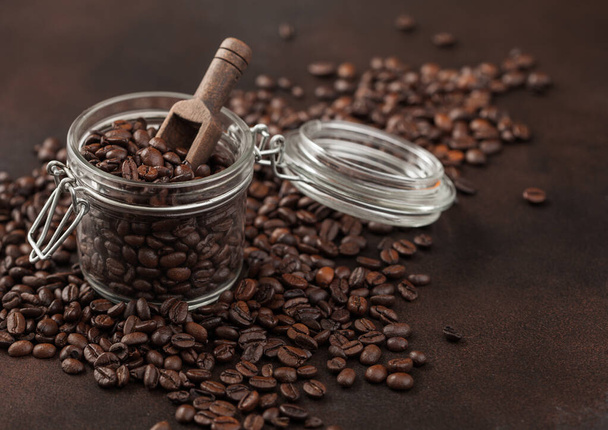 Roasted coffee beans in glass jar with wooden scoop on brown background. Macro - Foto, afbeelding