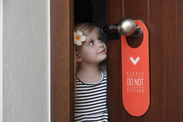 Rozkošná holčička v hotelovém pokoji otvírá dveře s prosím nerušte visačku visící na klice dveří. Koncept rodinné dovolené. - Fotografie, Obrázek