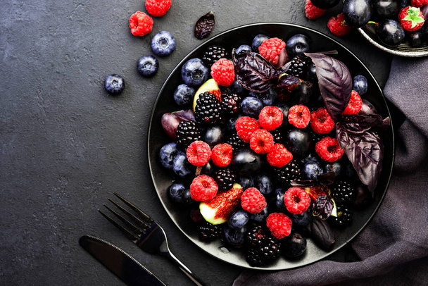 Autumn blue and black berries fruit vegan salad: blueberries, blackberries, grapes, figs and purple basil with honey dressing on dark background, top view - Photo, Image