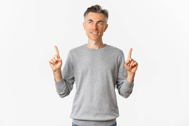 Image of handsome middle-aged man in grey sweater making his choice, smiling thoughtful, pointing fingers up and looking at product, standing over white background - Photo, Image