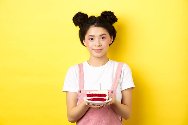Asiática cumpleaños chica de pie con pastel y sonriendo, celebrando b-day sobre fondo amarillo - Foto, imagen