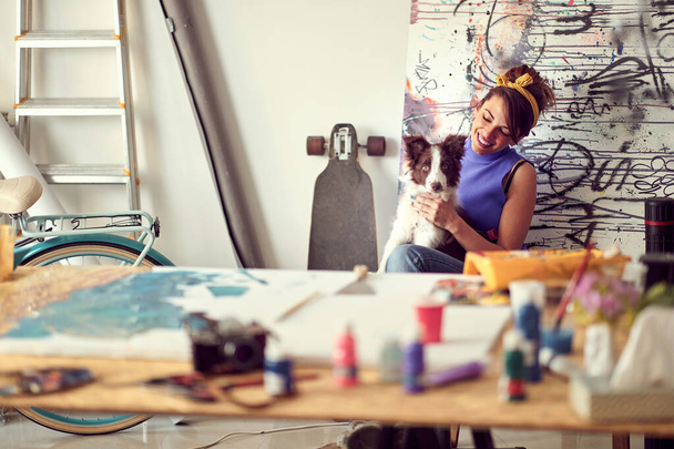 A young female artist is enjoying a break with her dog while working on a new painting in a pleasant atmosphere in the studio. Art, painting, friendship, studio - Photo, image