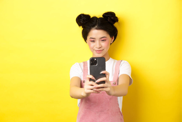 スマートフォンで写真を撮り、携帯電話でビデオを作り、笑顔で、黄色の背景に立っているスタイリッシュなアジアの女の子 - 写真・画像