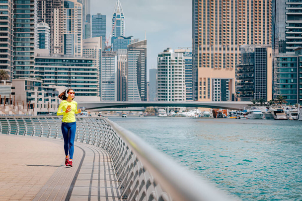 Mujer feliz con auriculares y gafas de sol trenes y hace correr y fitness en la acera del terraplén en el área de Dubai Marina - Foto, imagen