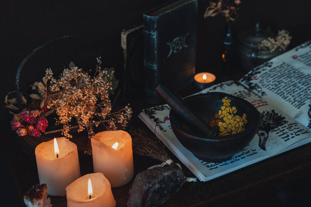 Wiccan witch altar prepared for casting a spell with an open Book of Shadows and pestle and mortar. Old spell books, crystals, burning lit candles, dried flowers in dark night background - Photo, Image