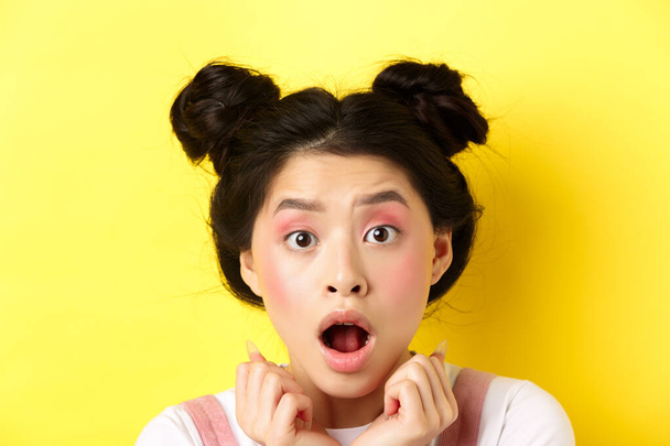 Head shot of shocked asian girl with hairbuns and glamour makeup, open mouth and looking startled at camera, standing against yellow background - Photo, Image