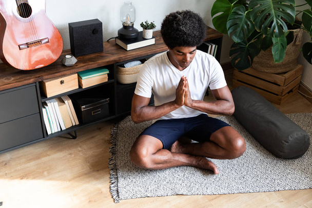Young african american man meditating with hands in prayer in bright living room. Meditation concept. Spirituality concept. - Photo, image