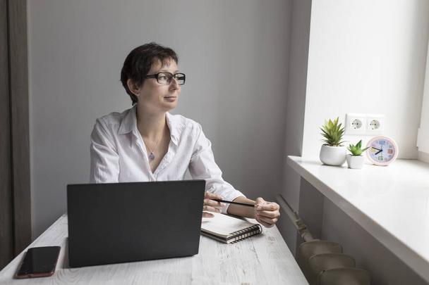 Middle-aged woman with short-haired brunette in glasses works at a laptop near window - Fotó, kép