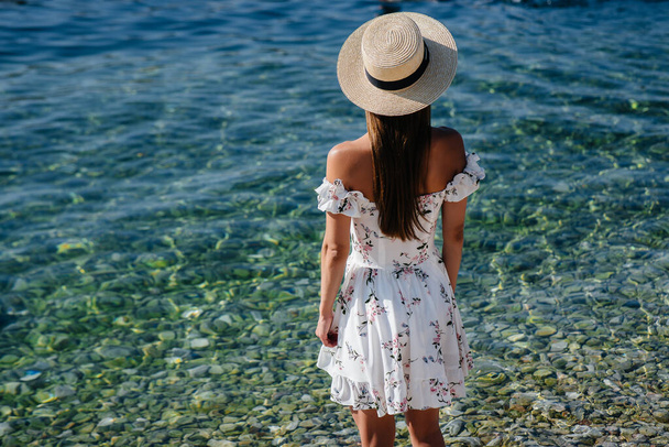 A beautiful young woman in a hat and a light dress with her back is walking along the ocean shore against the background of huge rocks on a sunny day. Tourism and vacation travel. - Fotoğraf, Görsel