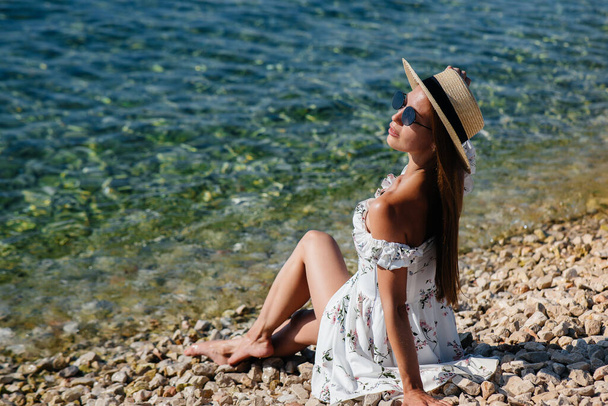 A beautiful young woman in a hat, glasses and a light dress is sitting on the ocean shore against the background of huge rocks on a sunny day. Tourism and tourist trips. - Photo, Image