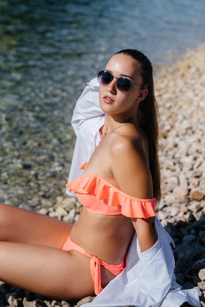 A beautiful young woman in glasses and a bikini is sitting on the ocean shore against the background of huge rocks on a sunny day. Tourism and tourist trips. - Foto, Bild